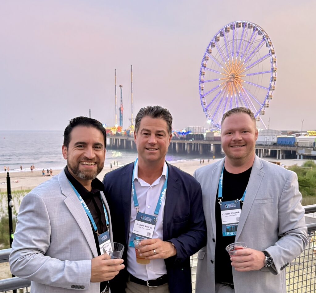 George Naveda, Aaron Metaj, and Curtis Taylor of Lima One Capital at NPLA Atlantic City conference smiling while standing in front of a boardwalk and Ferris wheel. 