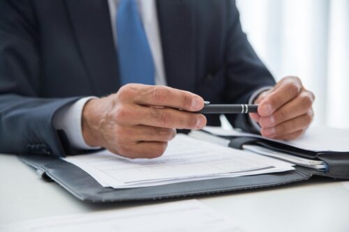 Man wearing suit and tie holding a pen above papers atop desk